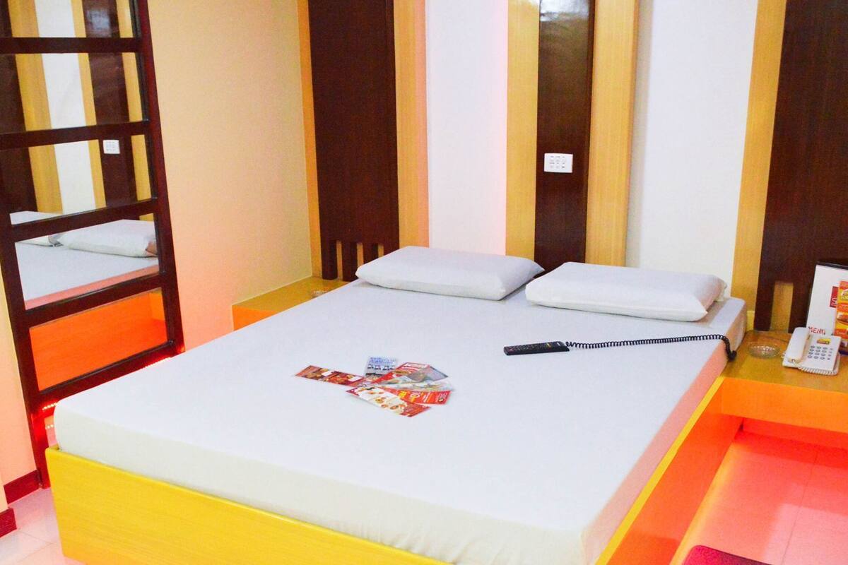 Una habitación de hotel con una cama, almohadas, un espejo, una mesita de noche con un teléfono y una revista.