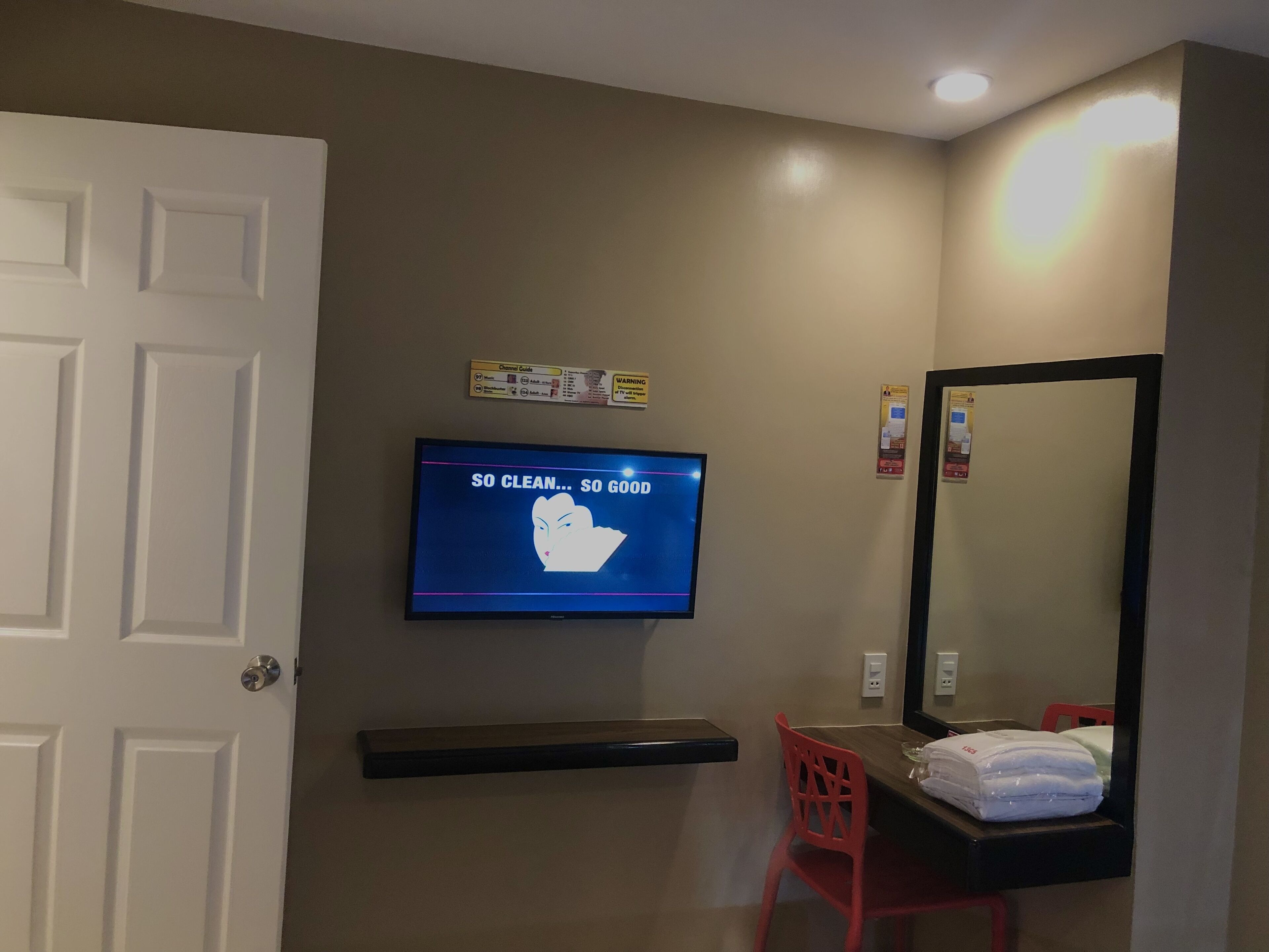 Habitación de hotel con una televisión mostrando un mensaje, un espejo, una silla roja y un perchero.