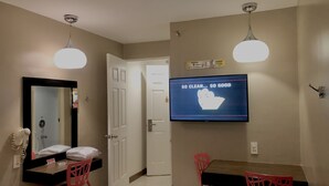 Habitación de hotel con televisor mostrando un mensaje de limpieza, un espejo, una puerta, un escritorio con dos sillas y un secador de pelo en un estante.