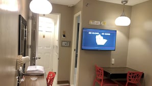 Habitación de hotel con una televisión mostrando un mensaje sobre la limpieza, una mesa de madera con una silla roja y una puerta blanca.