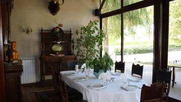 Un comedor con una mesa preparada para dos personas, un ventanal con vistas a la vegetación y un reloj fijado a la pared.