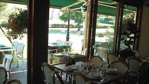 An indoor dining area with wicker chairs and tables, large windows with a view of the outdoors, and a green awning.