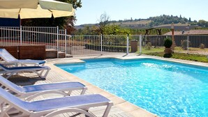 A rectangular swimming pool with clear blue water, surrounded by a white metal fence. There are several lounge chairs with blue cushions aligned along the poolside. A beige umbrella is partially visible in the upper left corner.