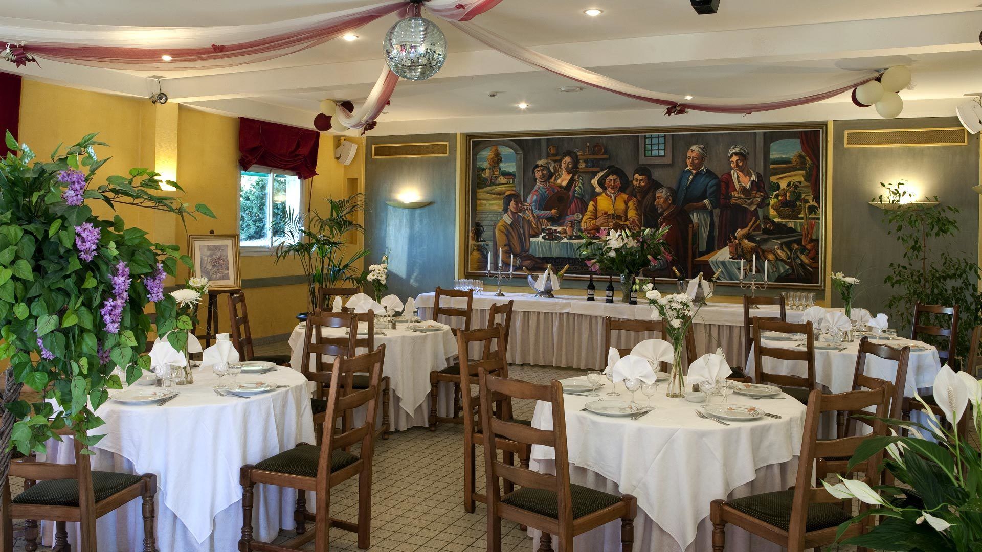 A restaurant with round tables, wooden chairs, and white tablecloths. There are potted plants and a large painting on the wall.
