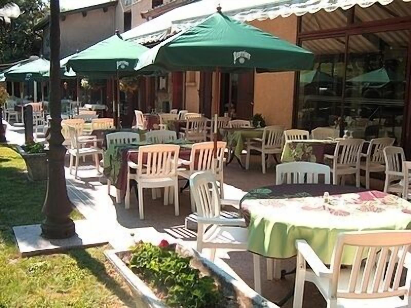 Ein Außenbereich mit Tischen und Stühlen, beschattet durch grüne Sonnenschirme, und ein Gebäude mit großen Fenstern im Hintergrund.
