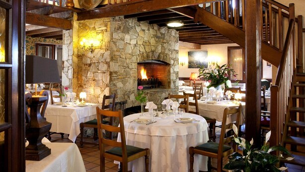 A restaurant with stone walls, wooden beams, and a fireplace. Tables are set with white tablecloths and chairs.