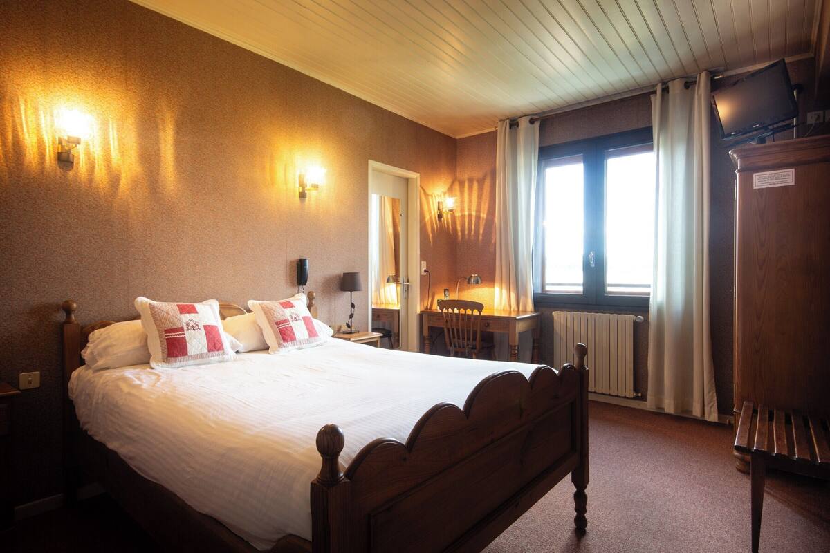 Een hotelkamer met een groot bed, een houten hoofdbord, een bureau en een stoel. Er is een raam met gordijnen, een radiator en een televisie op een standaard.