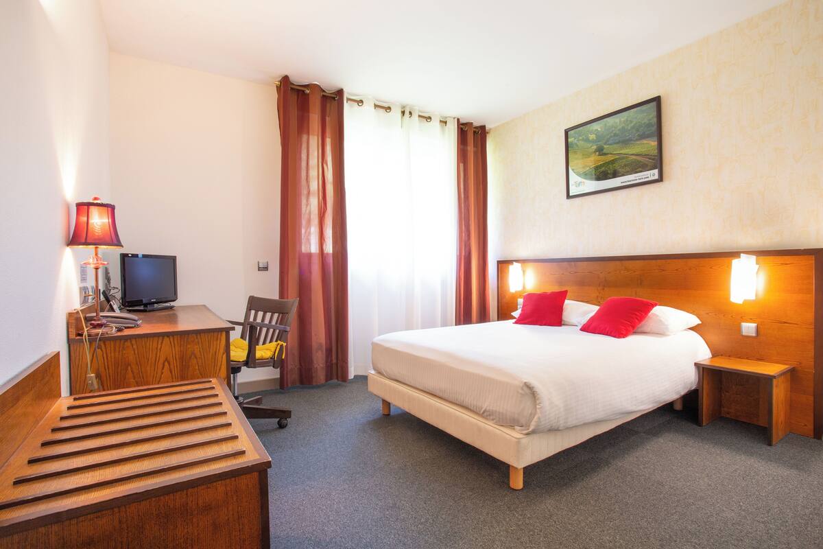 Een hotelkamer met een bed, een bureau met een computer, een stoel, een lamp en een schilderij aan de muur.