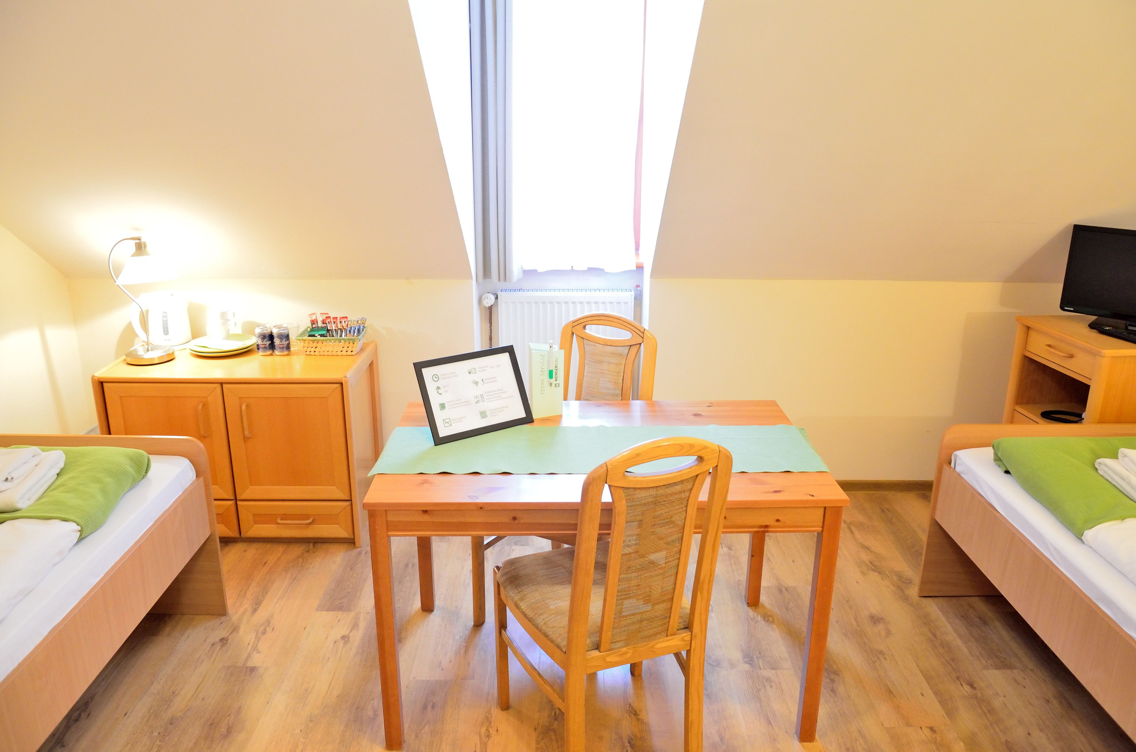 A room with a wooden desk, a chair, a bed with a green pillow, and a TV on a wooden stand.
