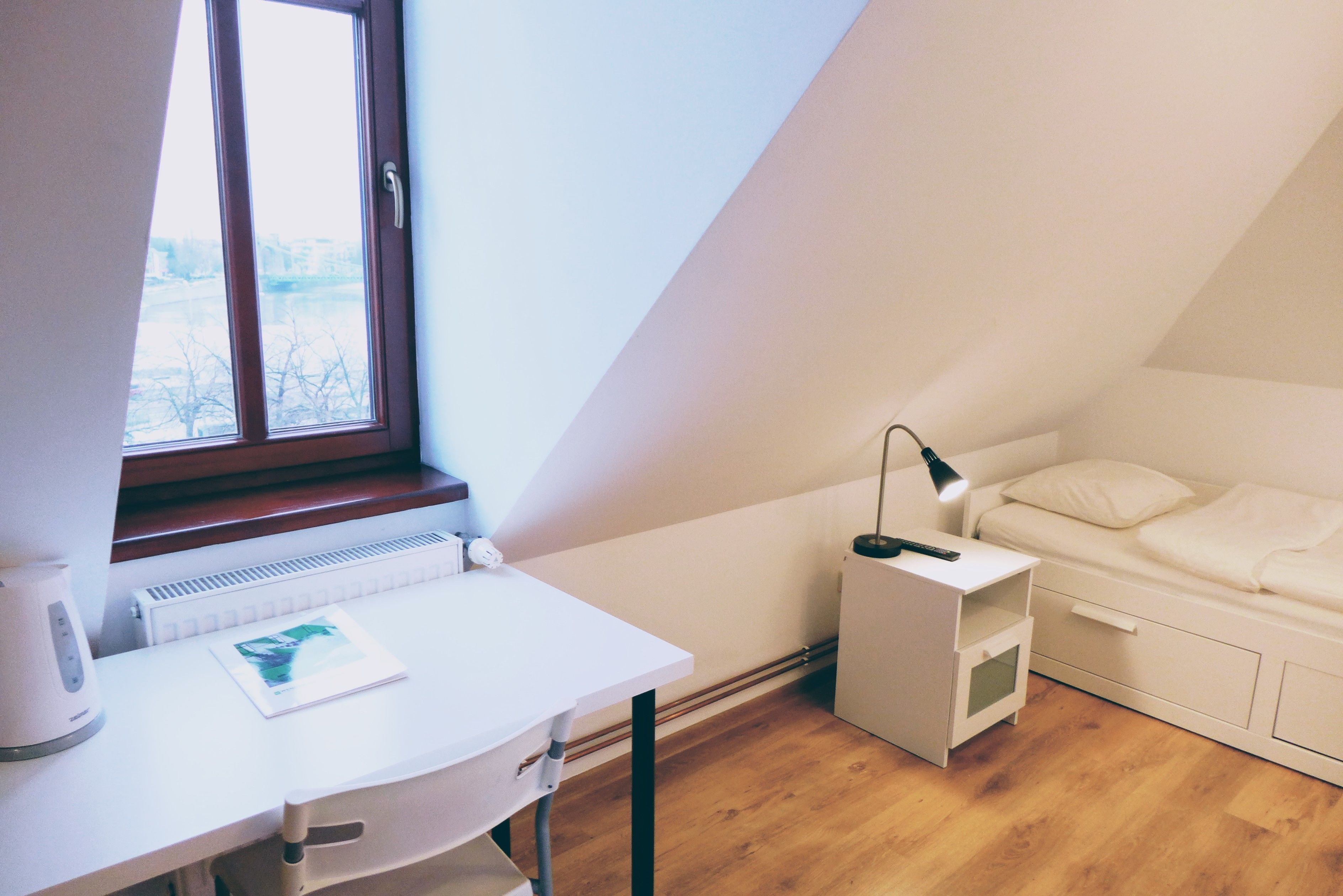 Um quarto pequeno estilo loft, com paredes brancas, teto inclinado, escrivaninha branca com luminária, radiador e janela com vista para árvores.