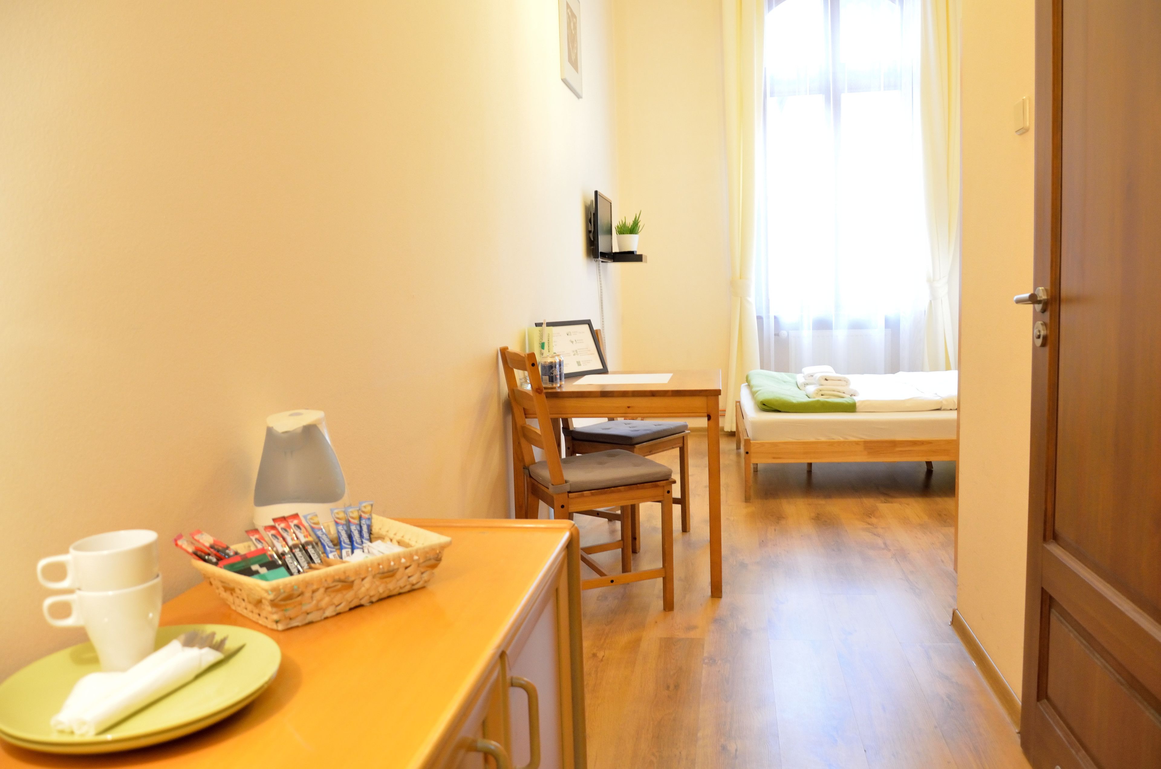 A small, cozy room with a wooden table, a chair, a bed with a green pillow, a bedside table with a lamp, and a window with sheer curtains.