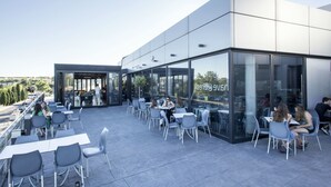 Un área de café al aire libre con mesas y sillas, un muro de vidrio con un letrero y la fachada de un edificio moderno.