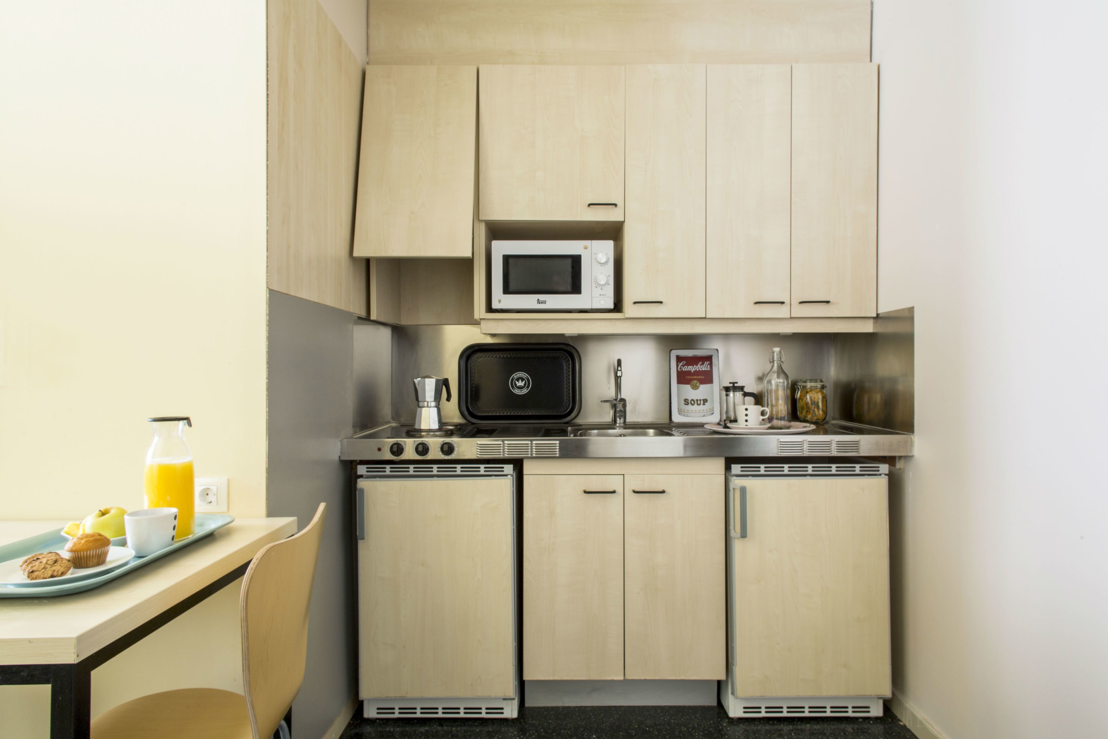 Een kleine keuken met een magnetron, kookplaat en spoelbak. Op het aanrecht staat een schaal met een muffin en een glas sinaasappelsap.
