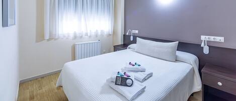 A hotel room with a large bed, a bedside table, a nightstand, a window with curtains, and a wall-mounted light.