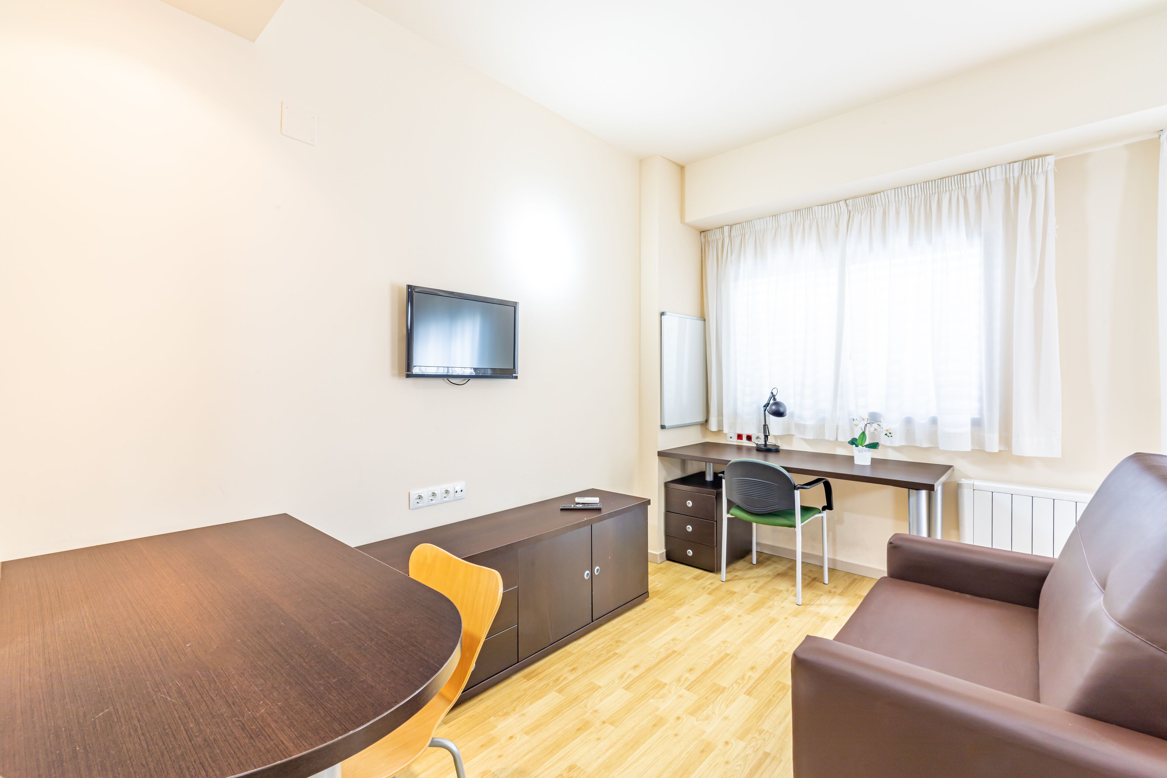 Une pièce moderne comprenant une table à manger en bois, un canapé en cuir marron, un bureau avec une chaise, un téléviseur et une fenêtre avec des rideaux.