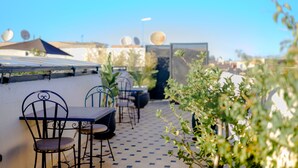 A rooftop patio with outdoor seating, including metal chairs and tables, surrounded by potted plants and a patterned floor.