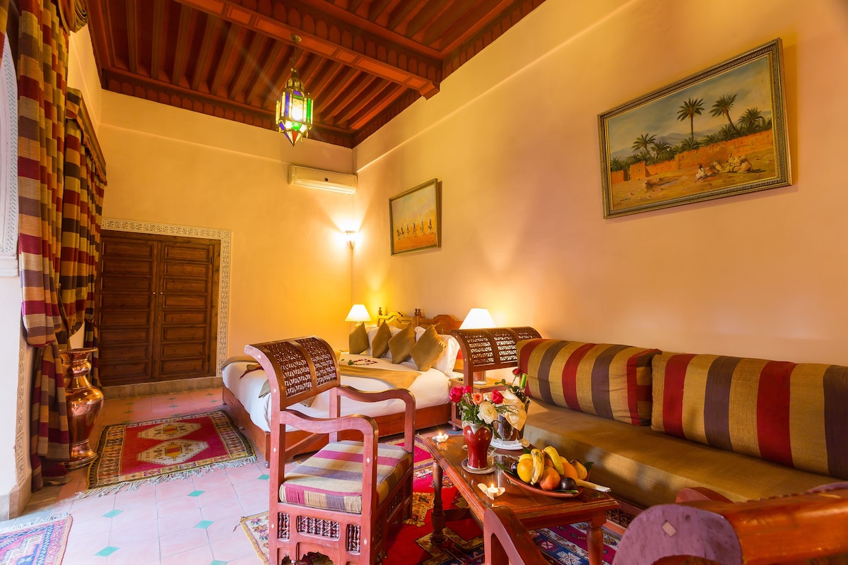 Una camera accogliente con soffitto in legno, un letto con cuscini, un divano, un tavolo con della frutta e un quadro appeso al muro.