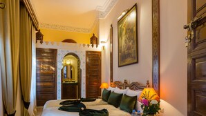 A traditional room with a large bed, a wooden side table with fruit, and a patterned rug.