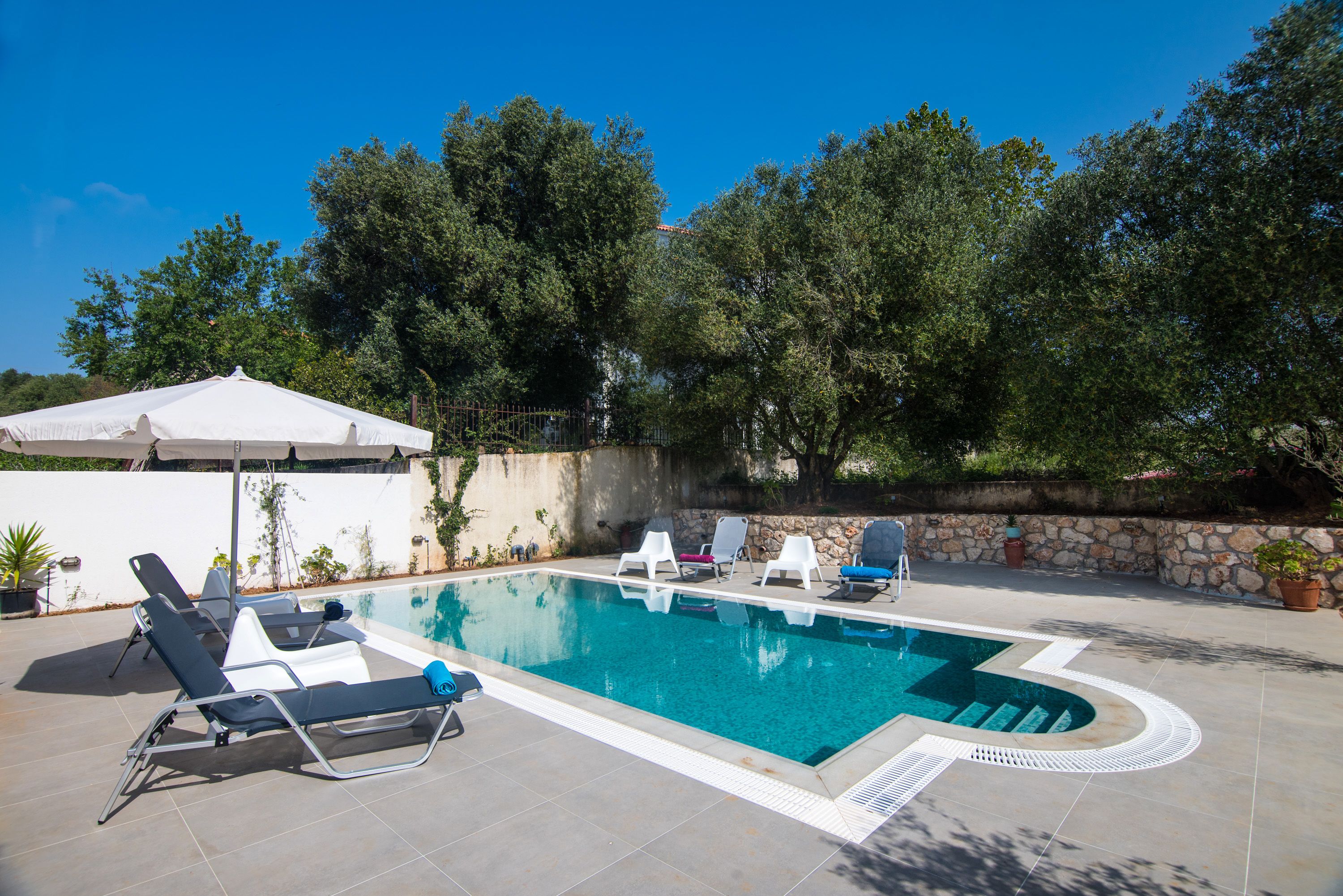 Piscina rectangular con solárium empedrado, rodeada de sillas de descanso y un paraguas blanco.