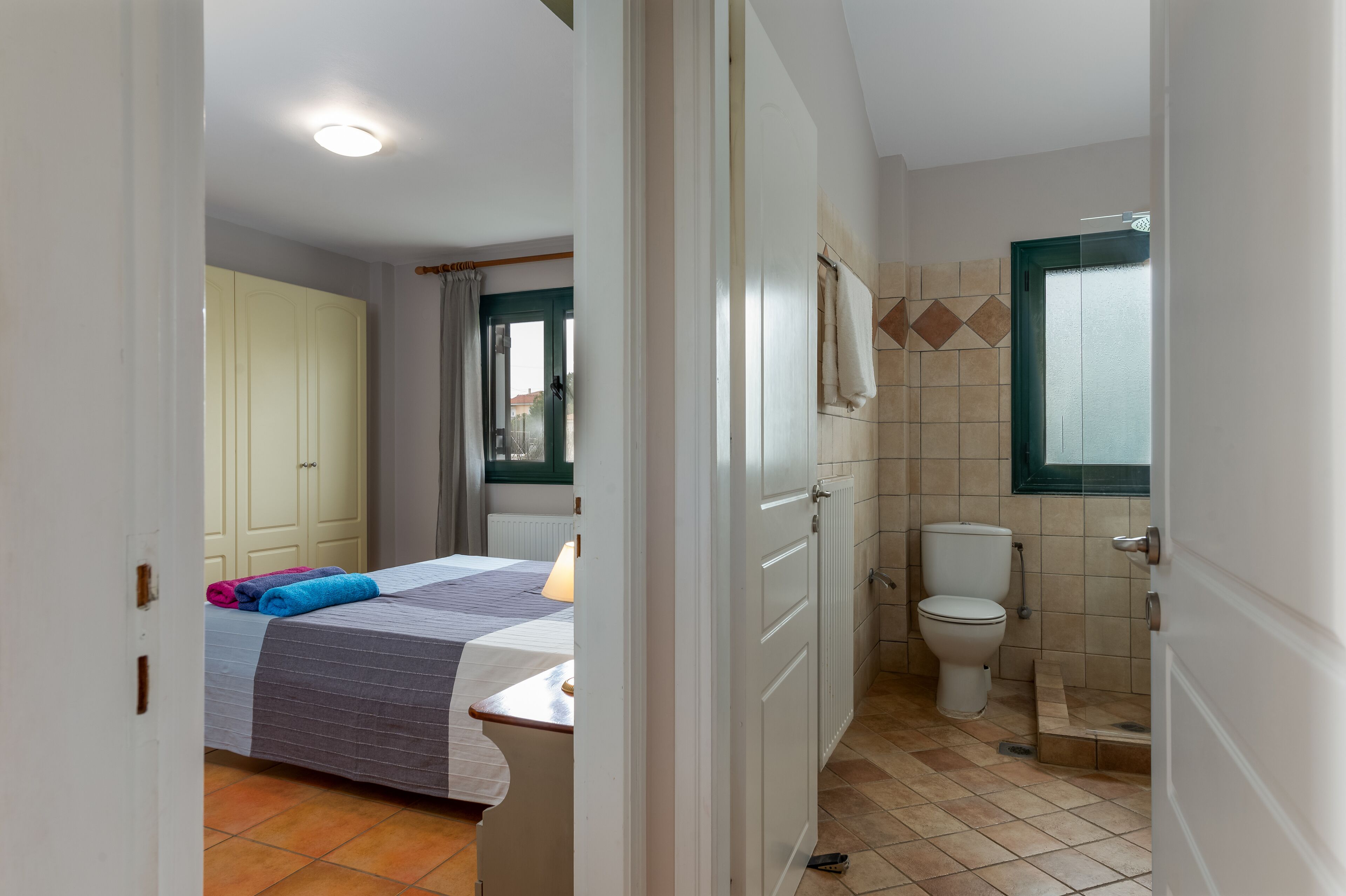 A bedroom with a bed, a bedside table, a window, a wardrobe, and a bathroom with a toilet and shower.