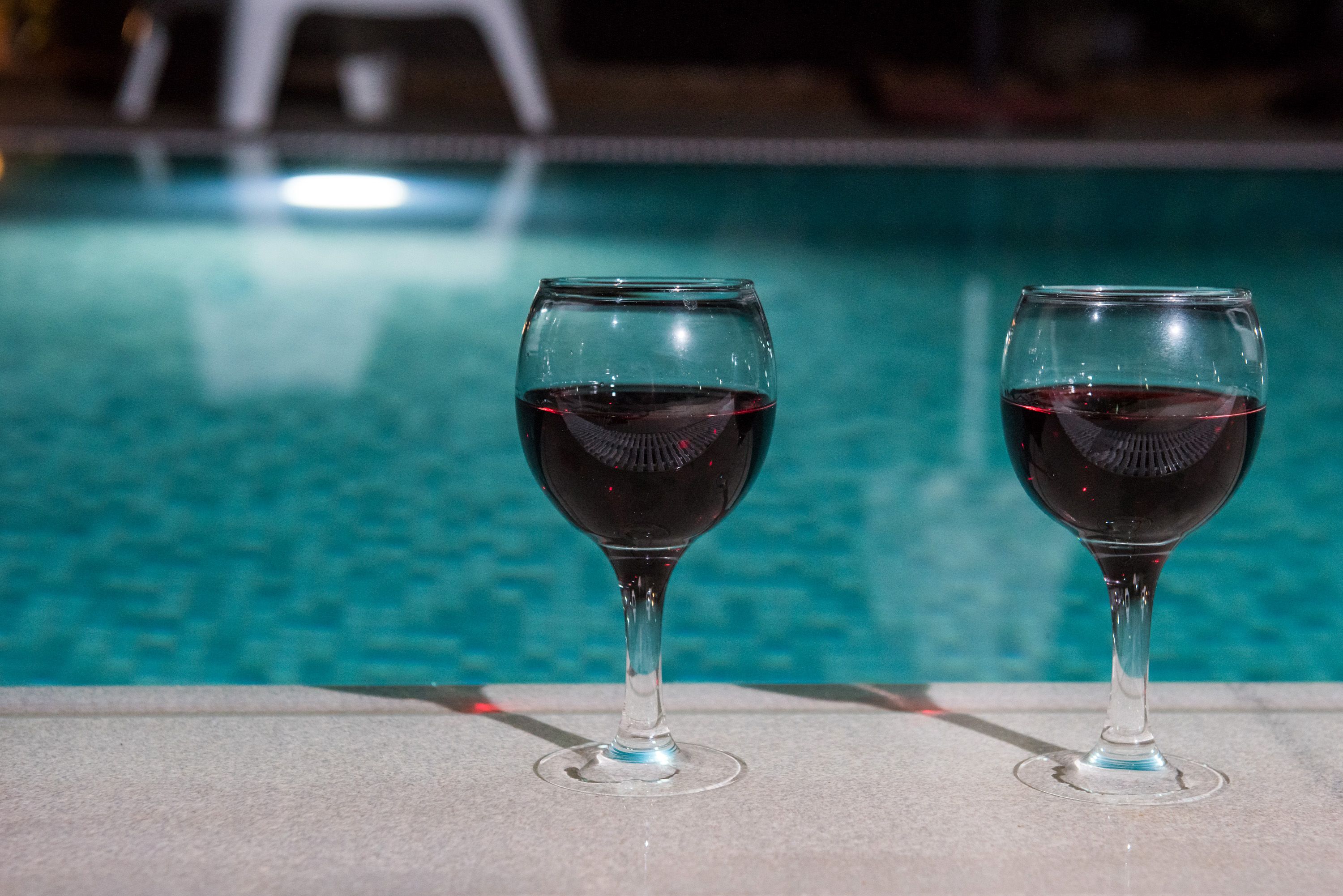Two wine glasses with red liquid on a poolside ledge.