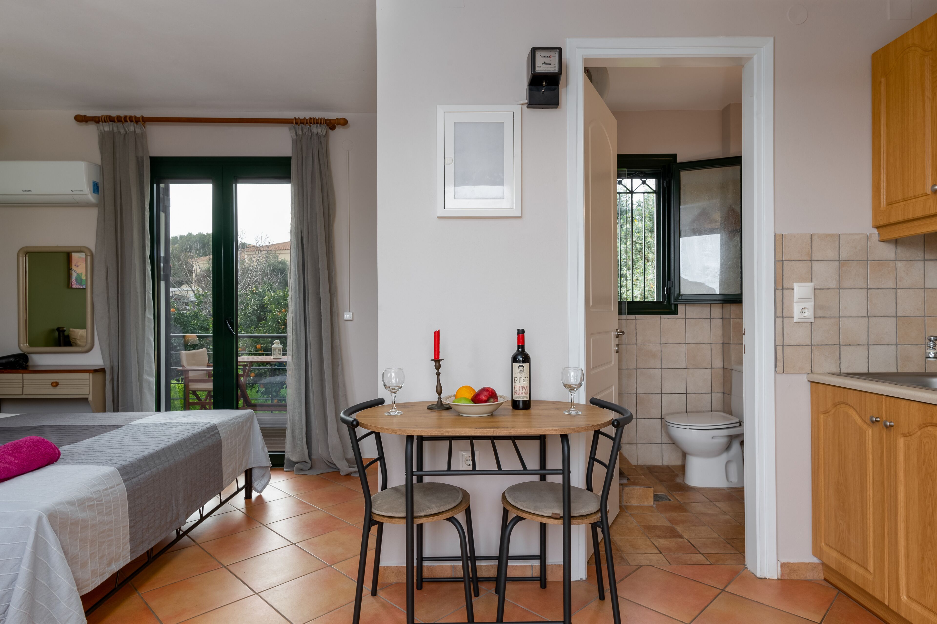 A room with a bed, a dining table with chairs, a small kitchen area, and a view through a door.