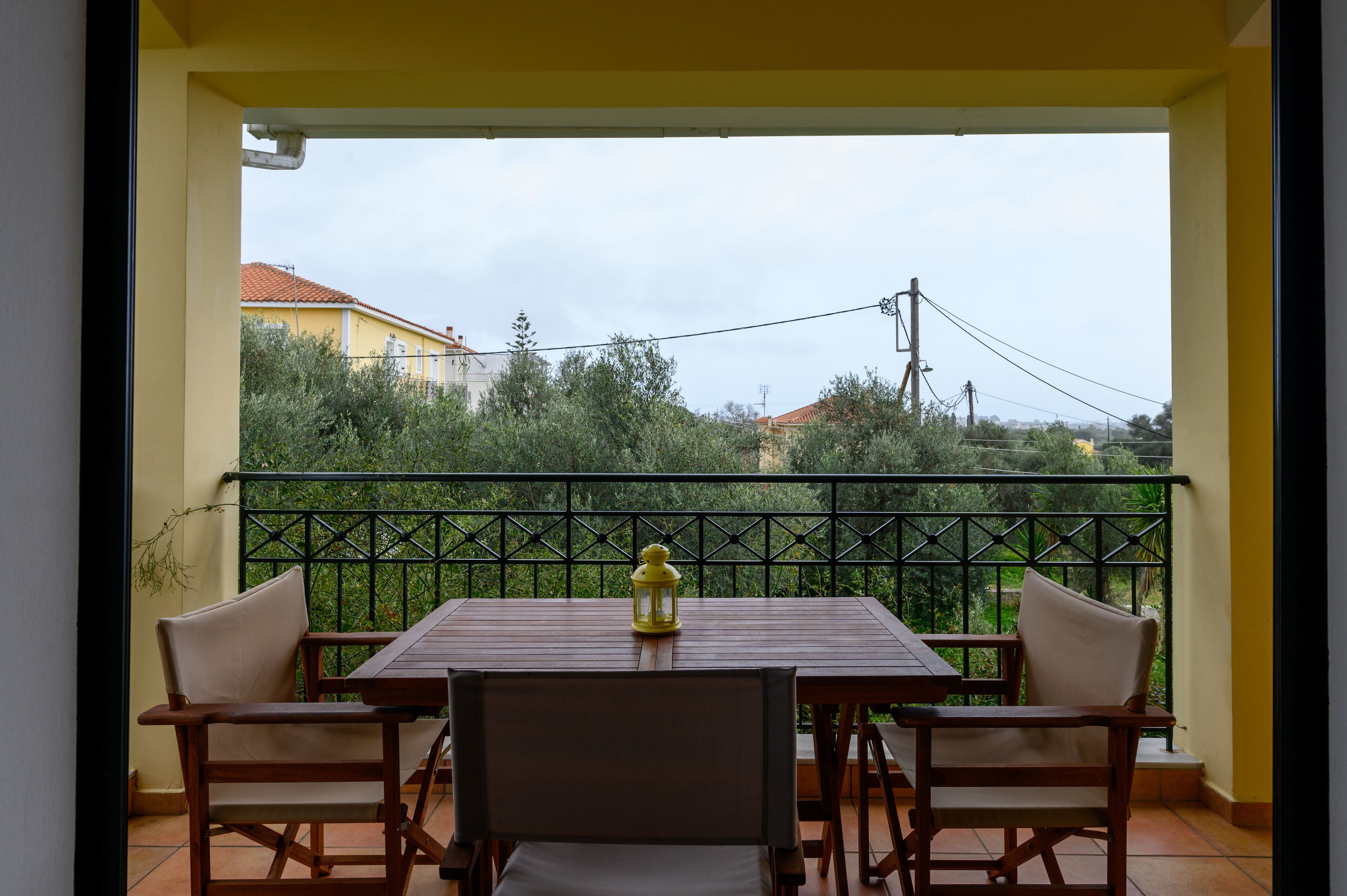 A balcony with a wooden table and two chairs, a yellow lantern, and a view of greenery and buildings.