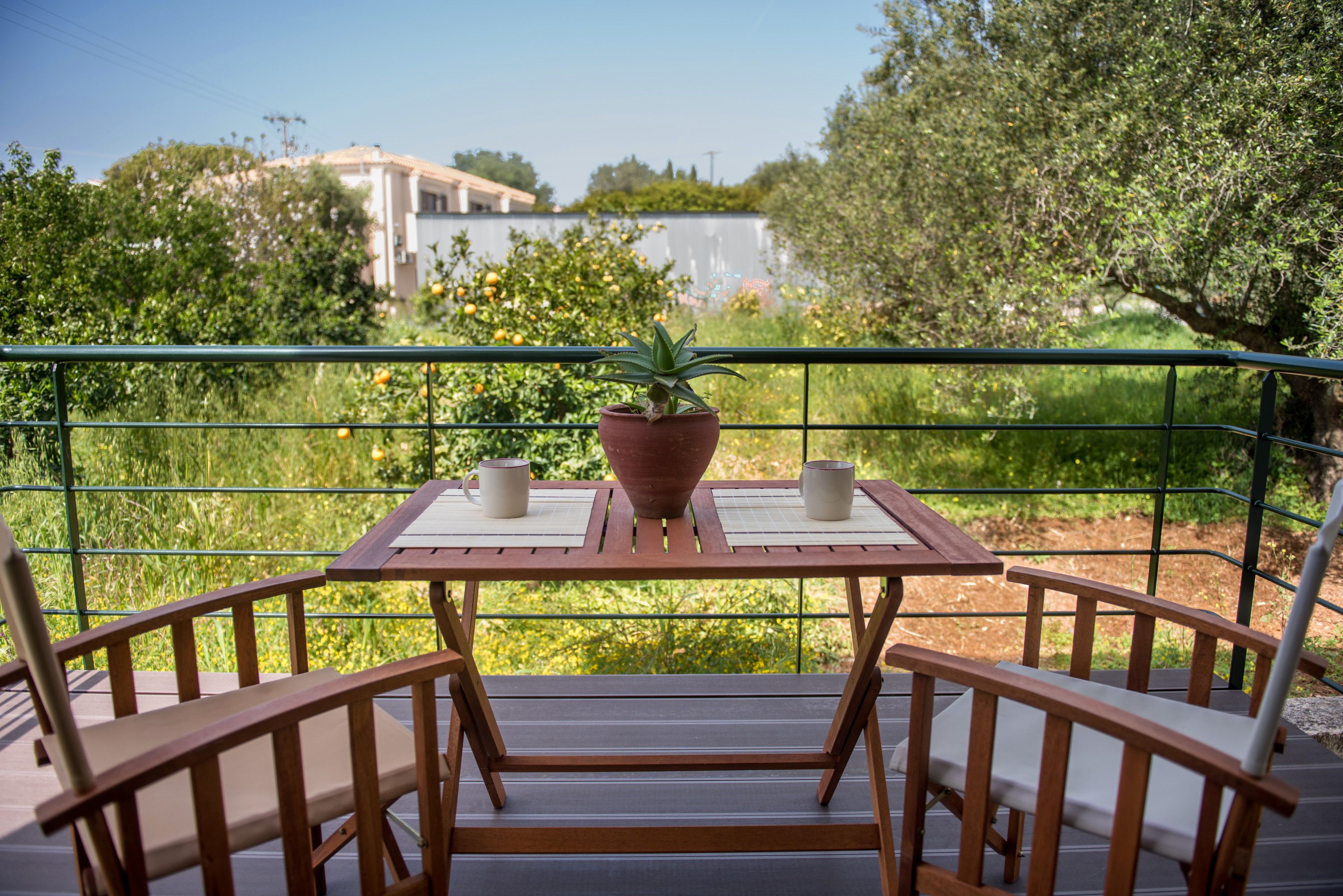 Een houten tafel met twee stoelen en een plant in pot, met uitzicht op een tuin met bomen en een gebouw op de achtergrond.
