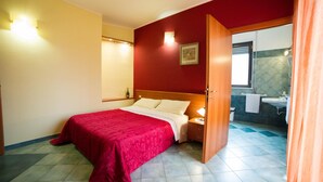 A hotel room with a bed, a nightstand, a wall-mounted shelf with a bottle and glasses, a painting, and a bathroom visible through an open door.