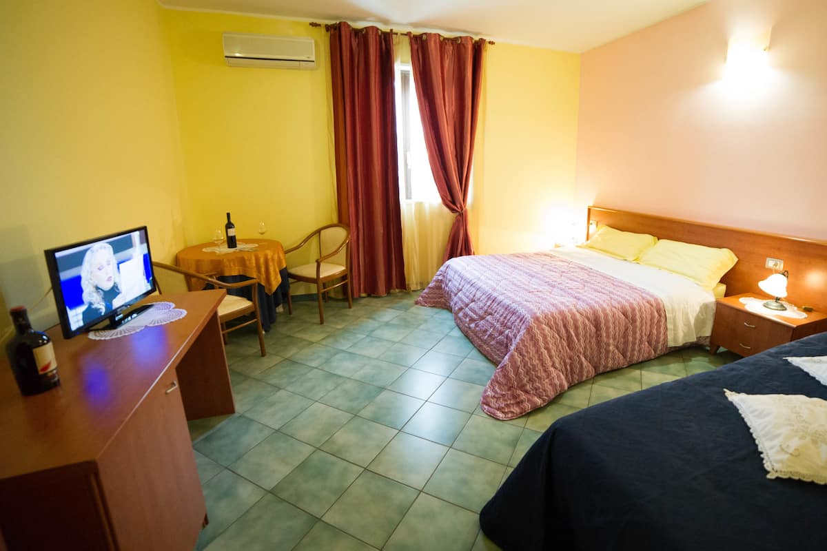 A hotel room with a bed, a desk with a TV, a chair, and a table with a bottle and glasses.