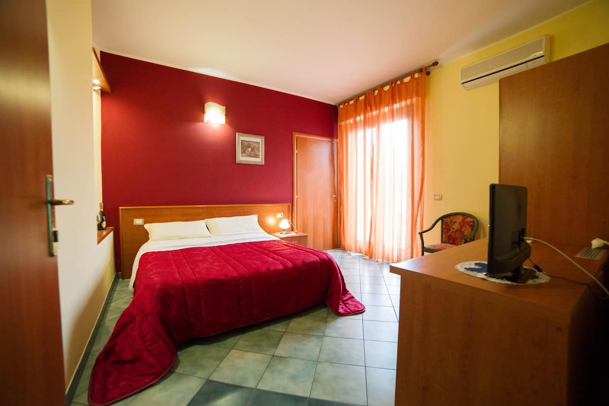 A hotel room with a bed, a desk with a computer, a chair, a nightstand, and a picture on the wall.