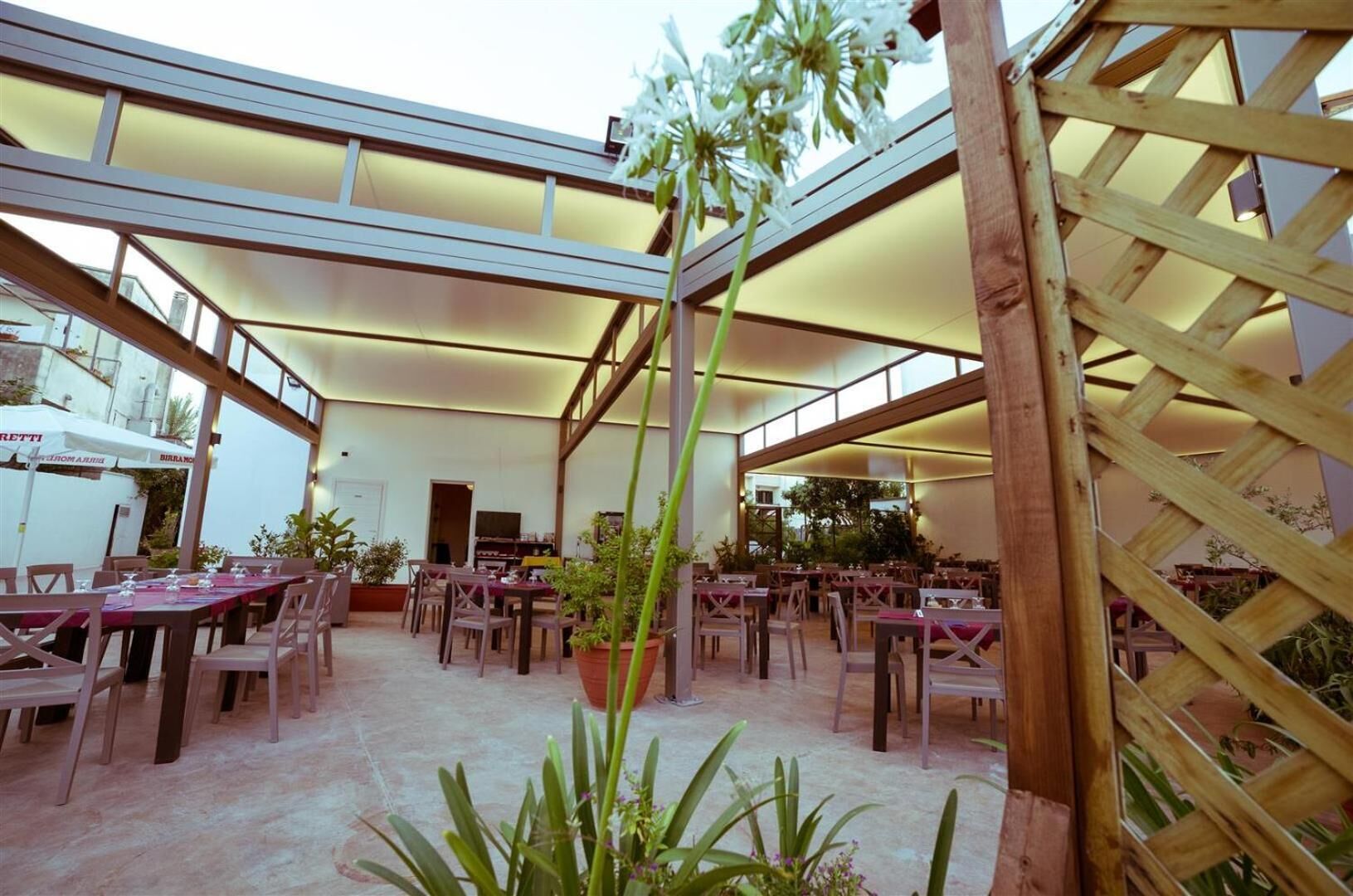 An outdoor dining area with multiple tables and chairs, surrounded by potted plants and wooden lattice structures.