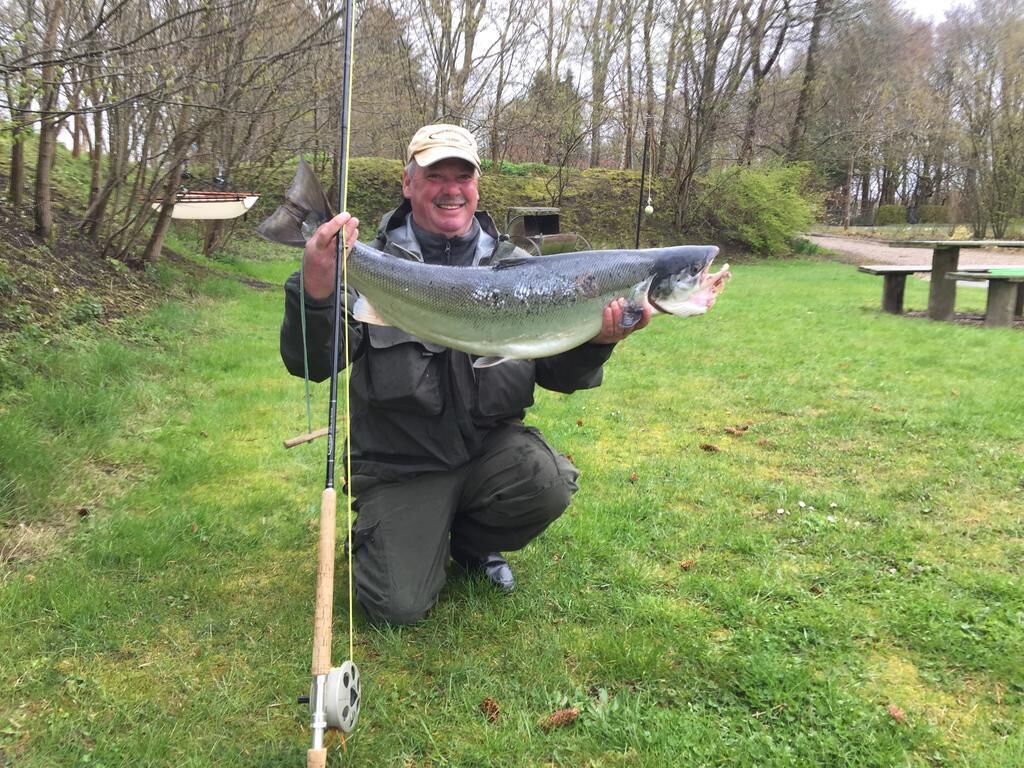 A person holding a large fish, standing in a grassy outdoor area with trees and a picnic table in the background.