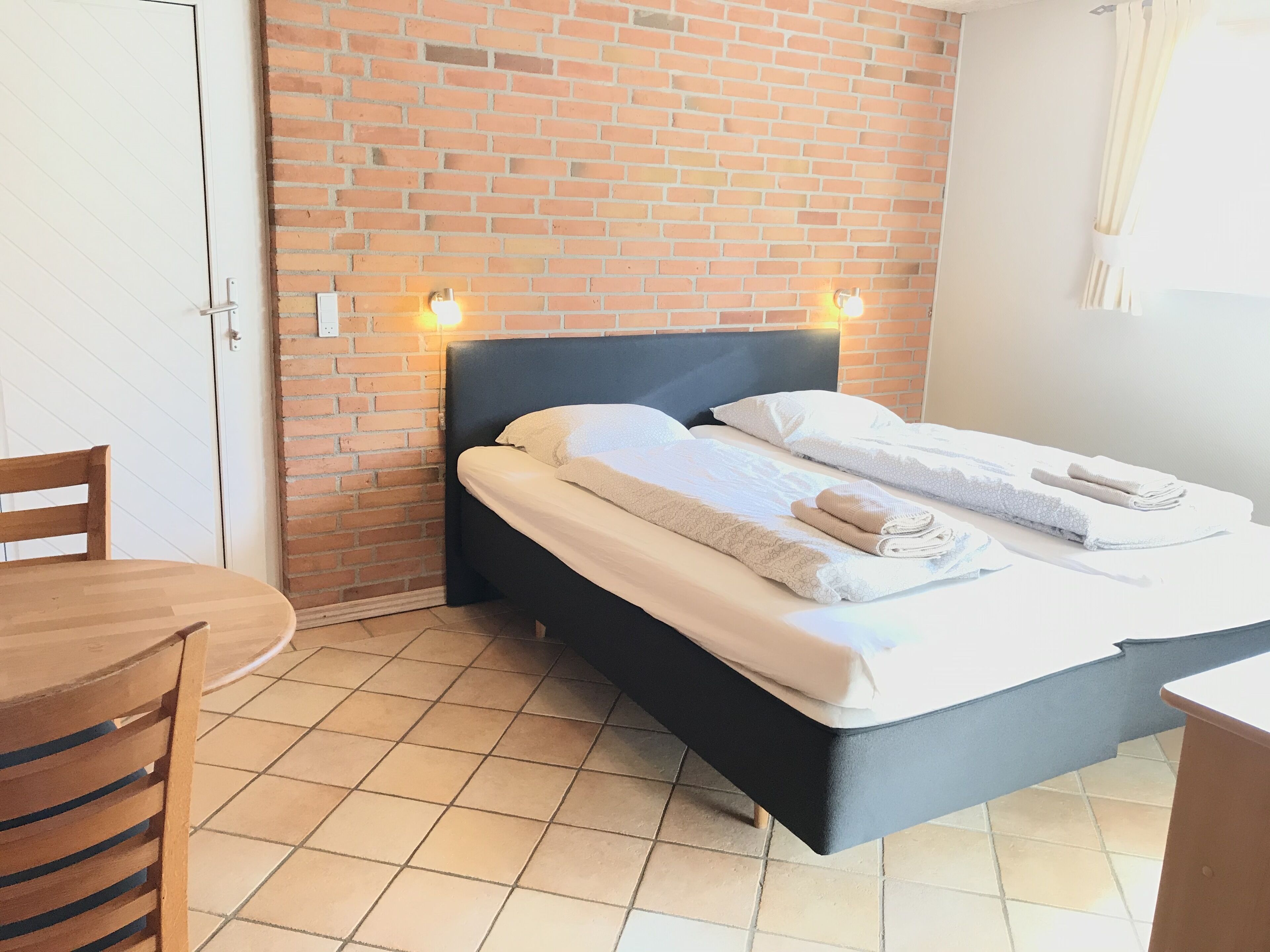 A room with a bed, a chair, a table, and a brick wall.