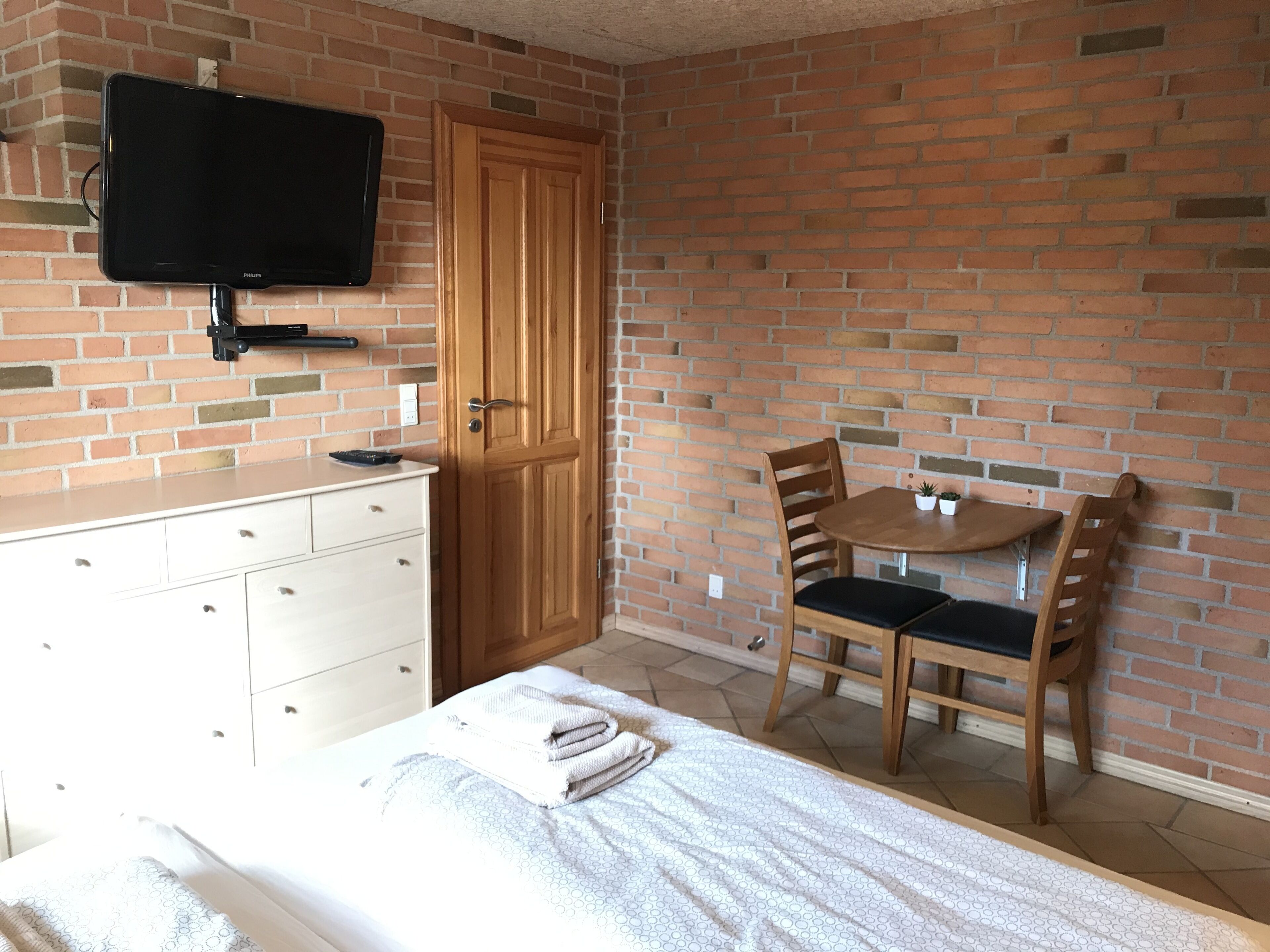 Ett hotellrum med en tegelvägg, en garderob i tr ä, en säng med ett vitt täcke, ett litet runt bord med två stolar och en platt-TV som är monterad på väggen.