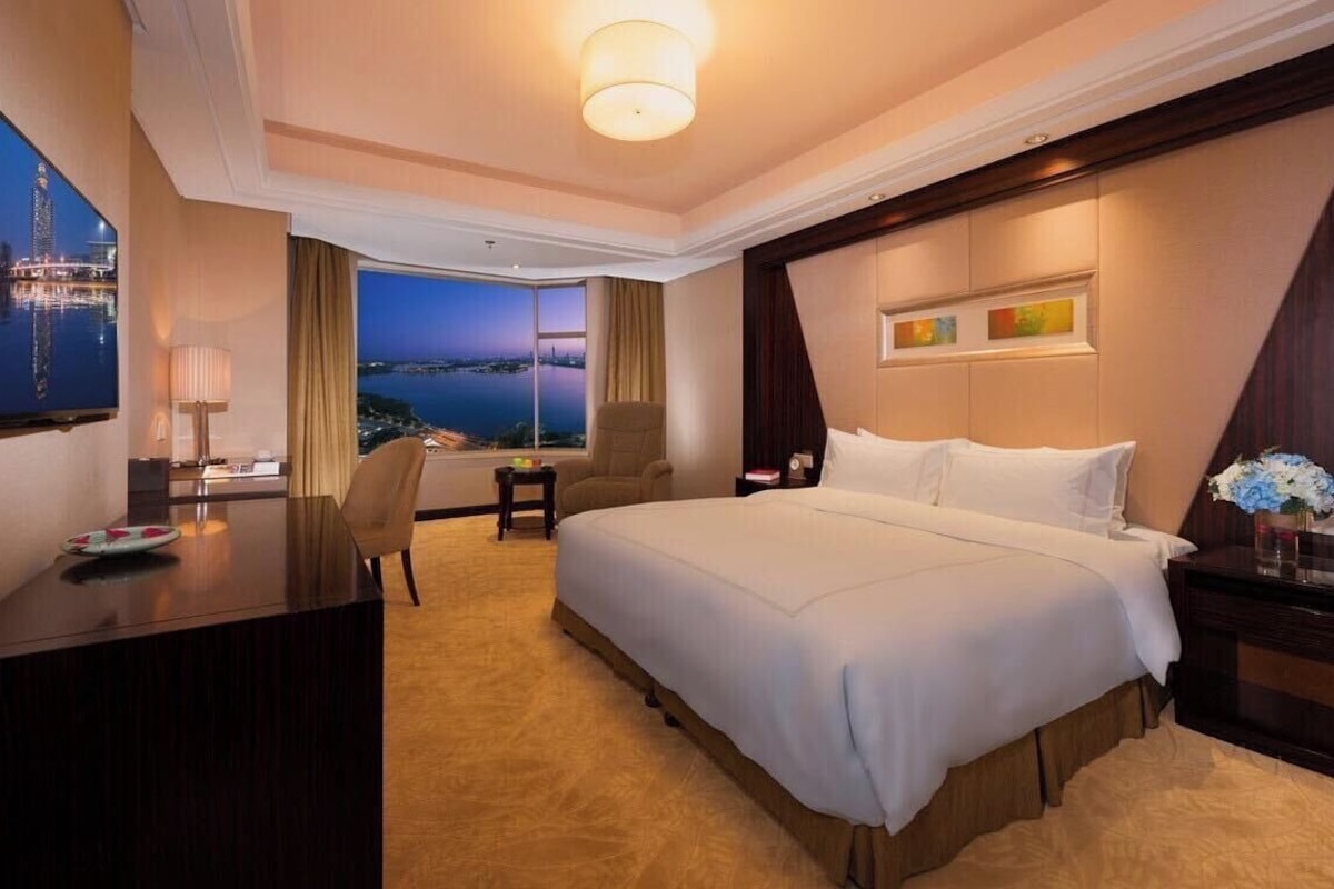 A hotel room with a large bed, a desk, a chair, a TV, and a view of a cityscape.