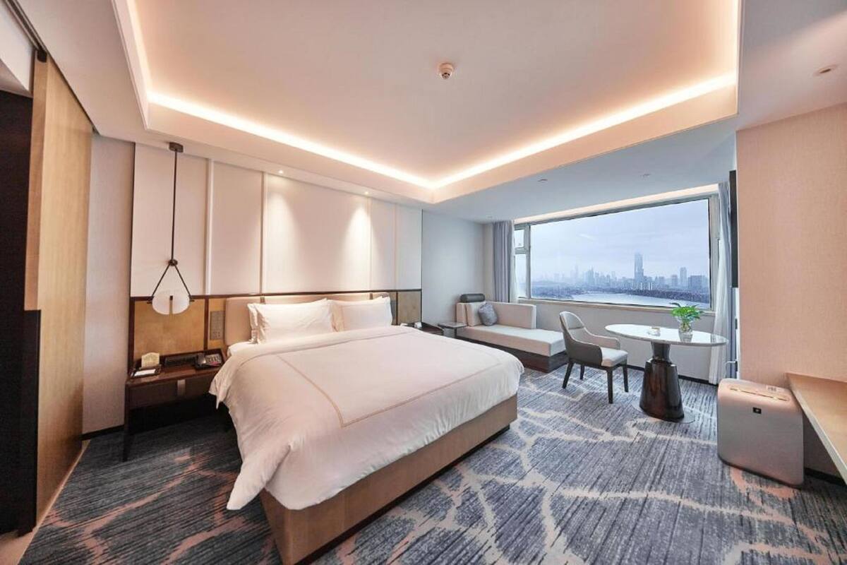 A modern hotel room with a large bed, a seating area, a small table, and a view of the city skyline.