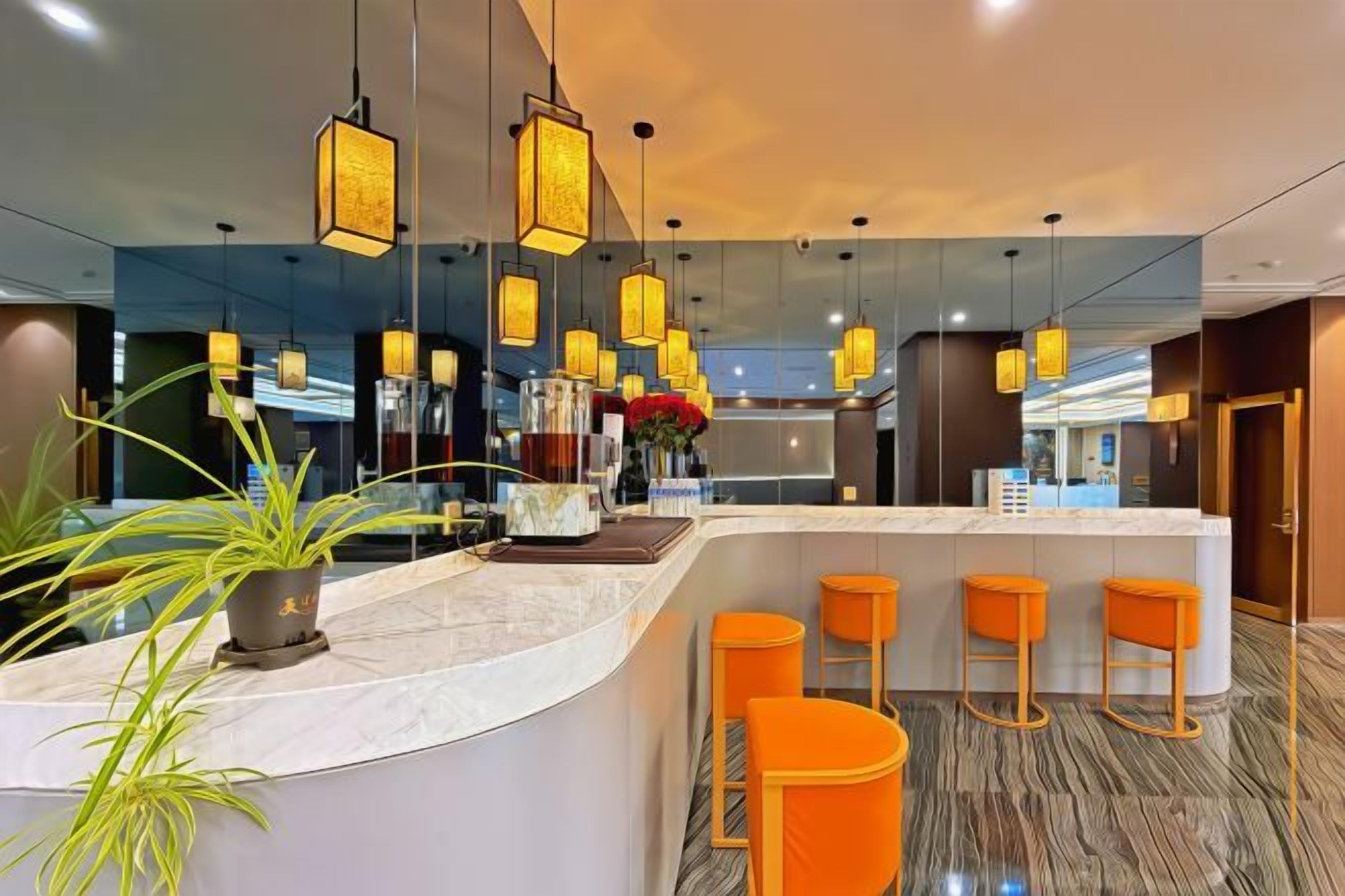 A modern hotel lobby with a marble reception desk, orange bar stools, hanging pendant lights, and a large mirror reflecting the space.