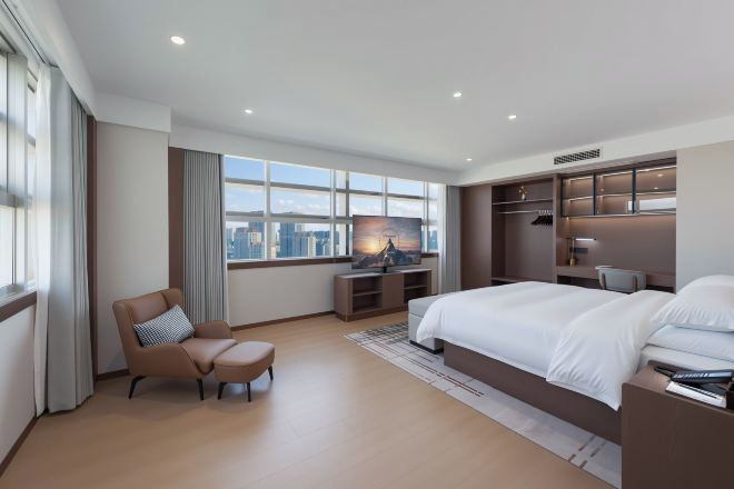 A modern hotel room with a large bed, a desk, a chair, and a view of the city.