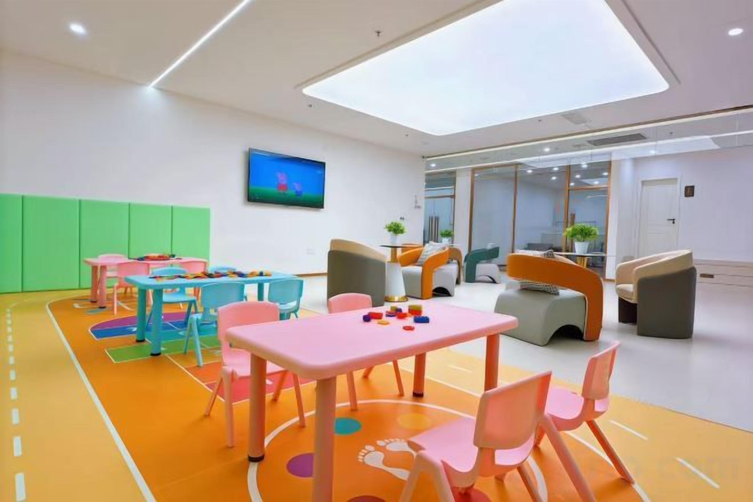 A brightly lit indoor space with tables and chairs arranged for activities, a large window, and a television on the wall.