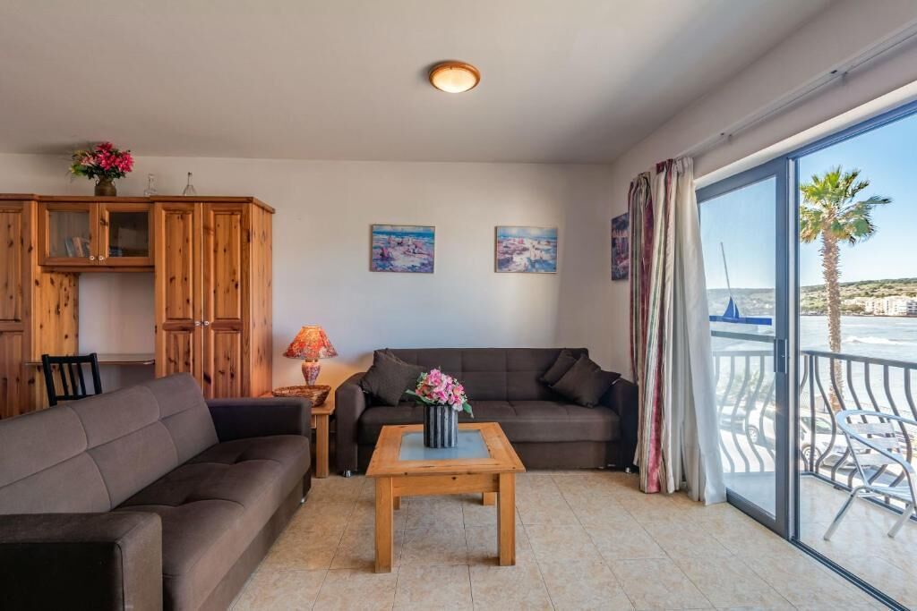 Een woonkamer met een bank, een houten kast, een bijzettafeltje met bloemen en uitzicht op zee vanuit een balkon.