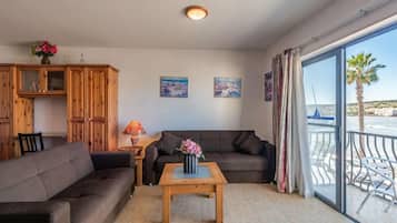 Een woonkamer met een bank, een houten kast, een bijzettafeltje met bloemen en uitzicht op zee vanuit een balkon.