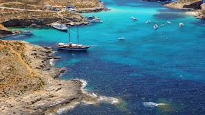 Une zone côtière où des bateaux sont amarrés près de rivages rocheux, avec des eaux turquoise et limpides, et une petite plage où se trouvent des gens.