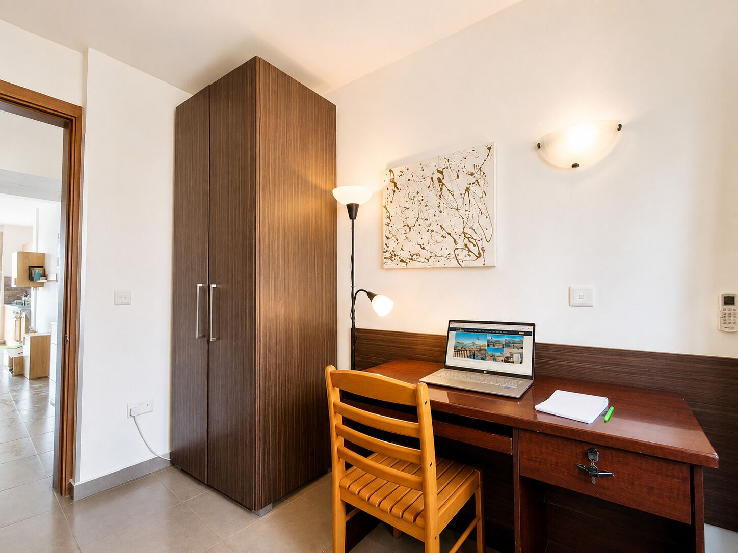 Ein Zimmer mit einem Holztisch, einem Laptop, einem Stuhl, einem Kleiderschrank und einer Wandleuchte.