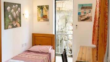 Un dormitorio con cama, escritorio y silla. Se ve una ventana con vista a un muro de piedra y un edificio.
