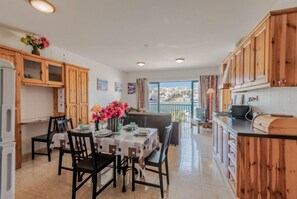 Uma sala de jantar com uma mesa posta para quatro pessoas, armários de madeira e vista pela janela.