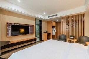 Una habitación de hotel moderna con una cama grande, un televisor montado en la pared, una zona de estar con sillas y una mesita con una planta.