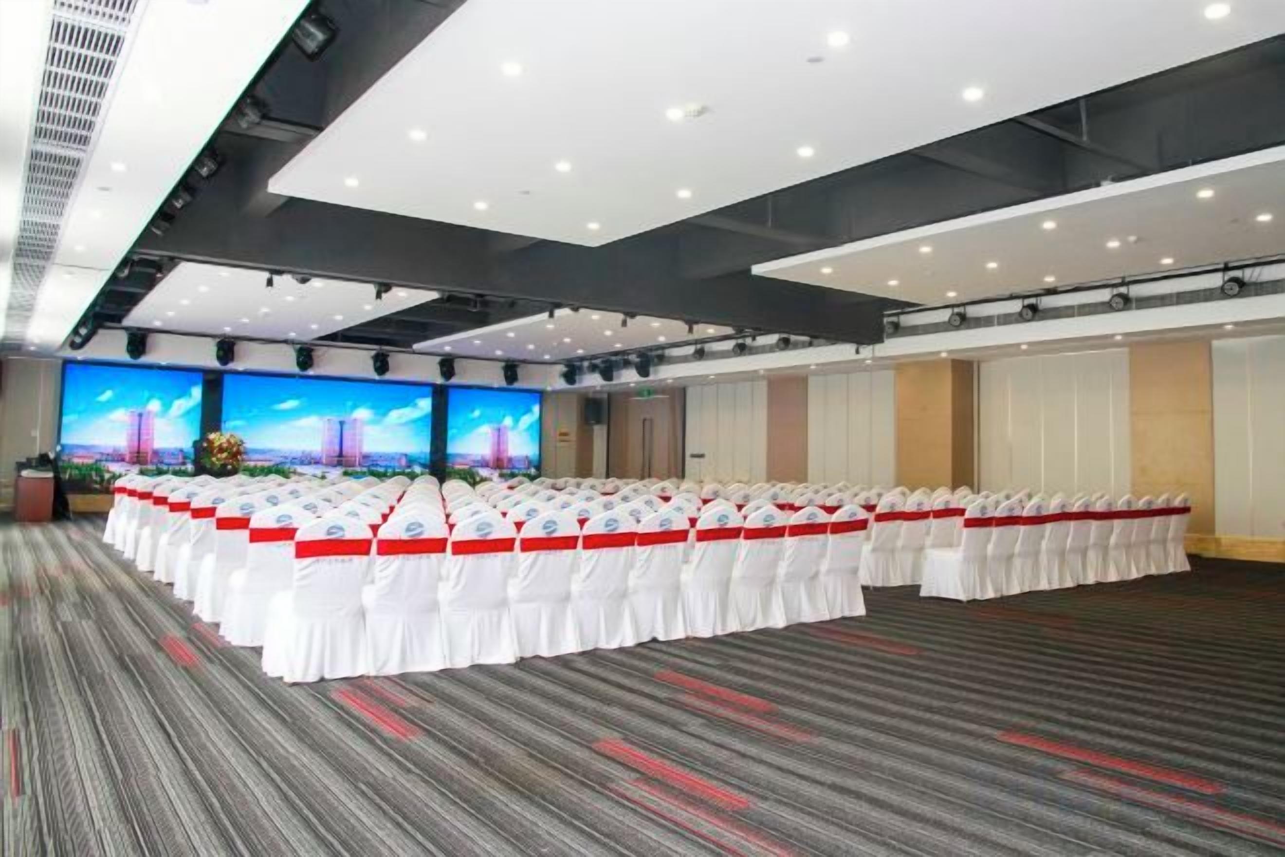 Un salón de conferencias con filas de sillas blancas, una pantalla grande y piso alfombrado.