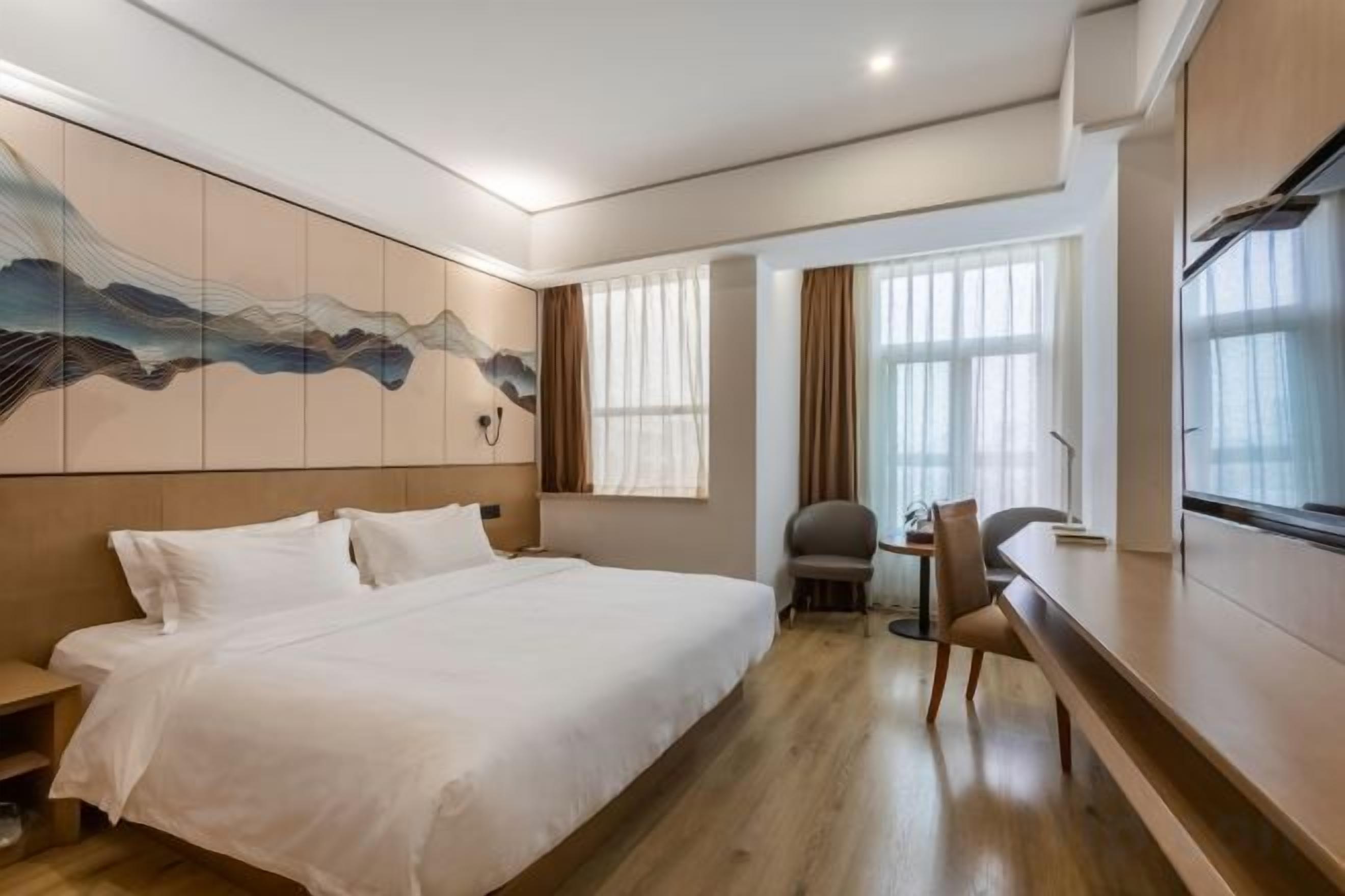 A hotel room with a large bed, a desk, and a chair. There is a window with curtains, a wall-mounted artwork, and a wooden floor.