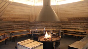 Een rustiek houten interieur met een open haard in het midden, omringd door banken en tafels.